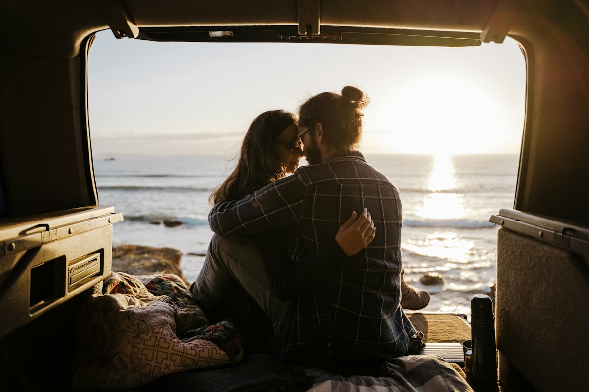 Two travellers sitting in the open back of a van, looking out at the sea at sunset