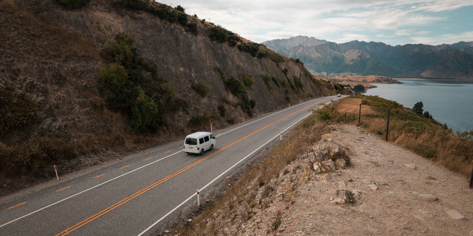 A white campervan driving along a lakeside road below mountains in New Zealand