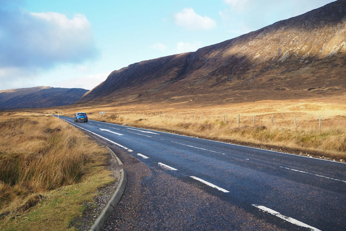 An empty road crossing open moorland toward mountains in the Scottish Highlands